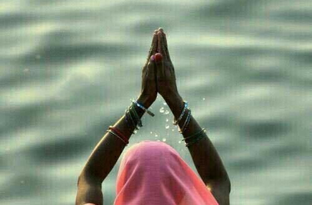 A woman in a pink sari with her hands raised in prayer at the water.